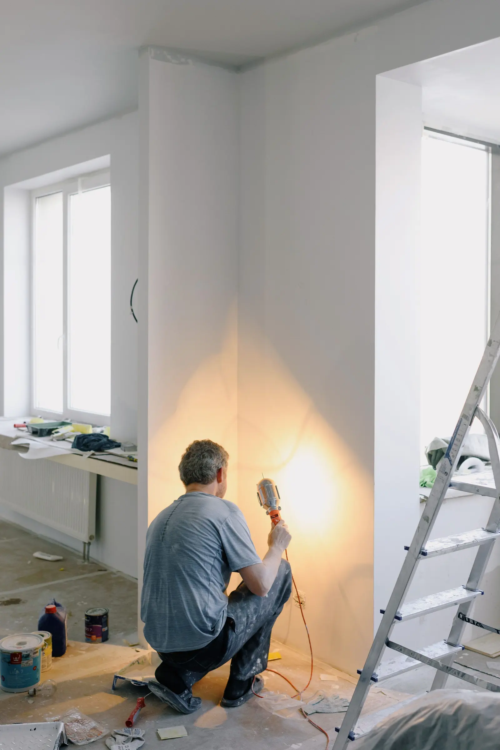Dans un appartement en travaux, un artisan agenouillé éclaire le mur avec sa lampe de chantier.